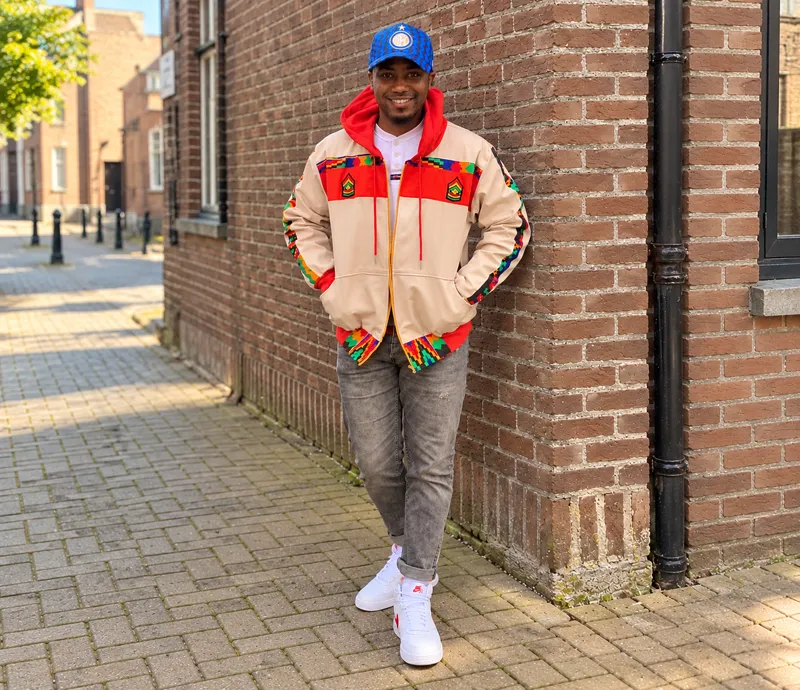 Customer in a beige and red print bomber jacket and blue cap on a brick street